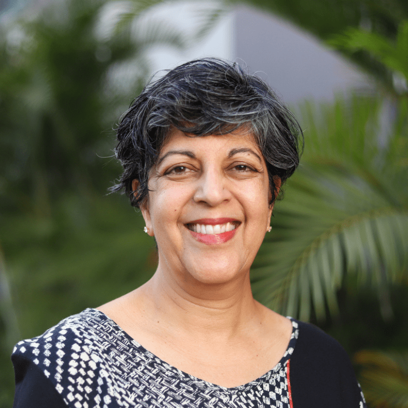 Anu Kumar's headshot - woman with short dark hair wearing a black and white patterned top with blurred foliage in the background