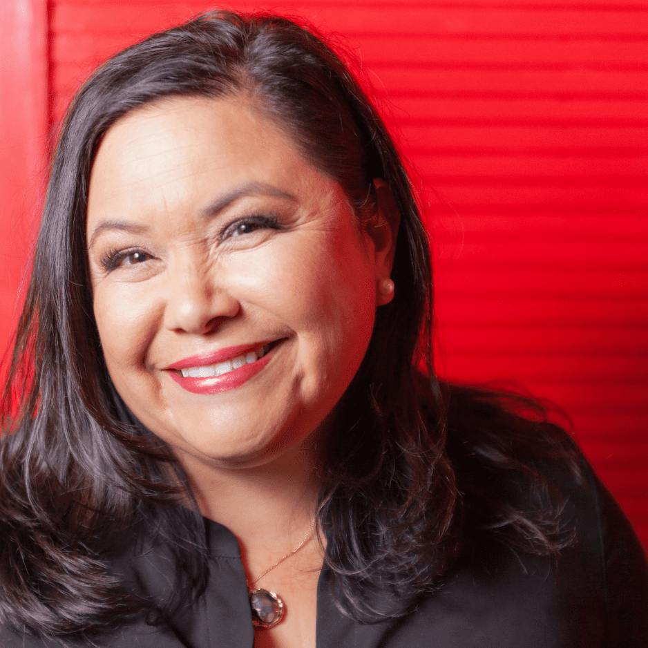 Picture of Theresa Gonzales who has brunette hair past her shoulders, wearing a black collared shirt, necklace, and red lipstick in front of a red slatted background