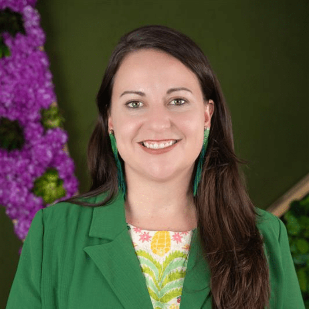 Verónica Colón-Rosario's headshot - woman with long, brown hair wearing a green blazer with a colorful top underneath and green parrot earrings